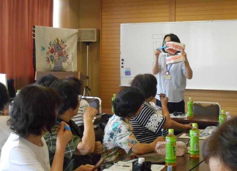 ホワイトボードがついた壁の前に立ち、大きな歯の模型に歯ブラシを当てている女性と、席に座り同じように歯ブラシを自分たちの歯にあてている女性たちの写真