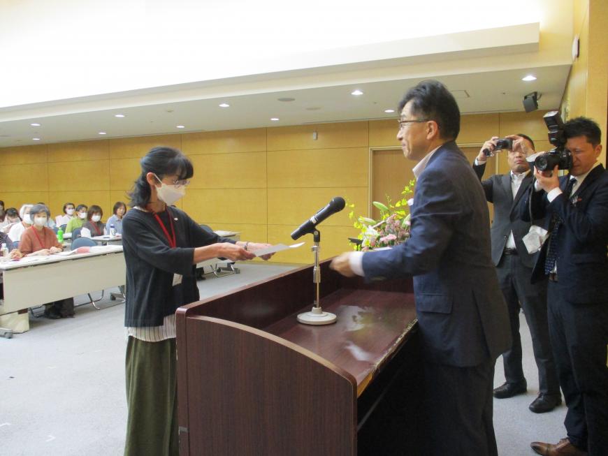 出席者たちが長机に並んだ席に座る中、マイクの置かれた演台の後ろに立った市長が演台を挟み向かい合って立っている女性に書状を渡している写真