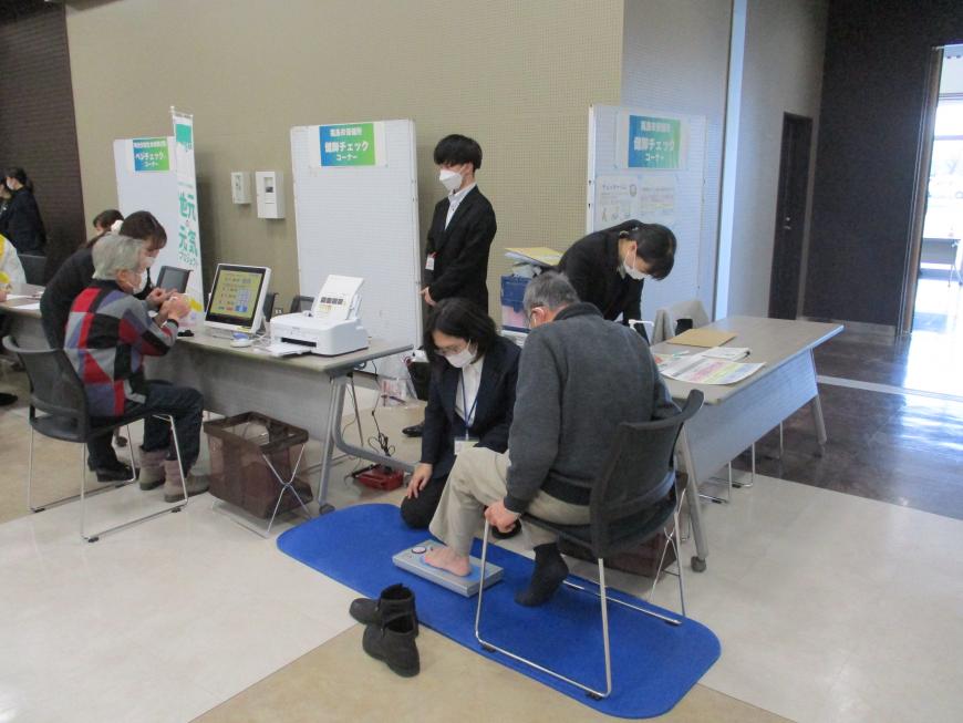 壁沿いに設置されたテーブルの上や床の青いマットの上に置かれた機器を、スタッフのサポートを受けながら来場者が試している様子の写真