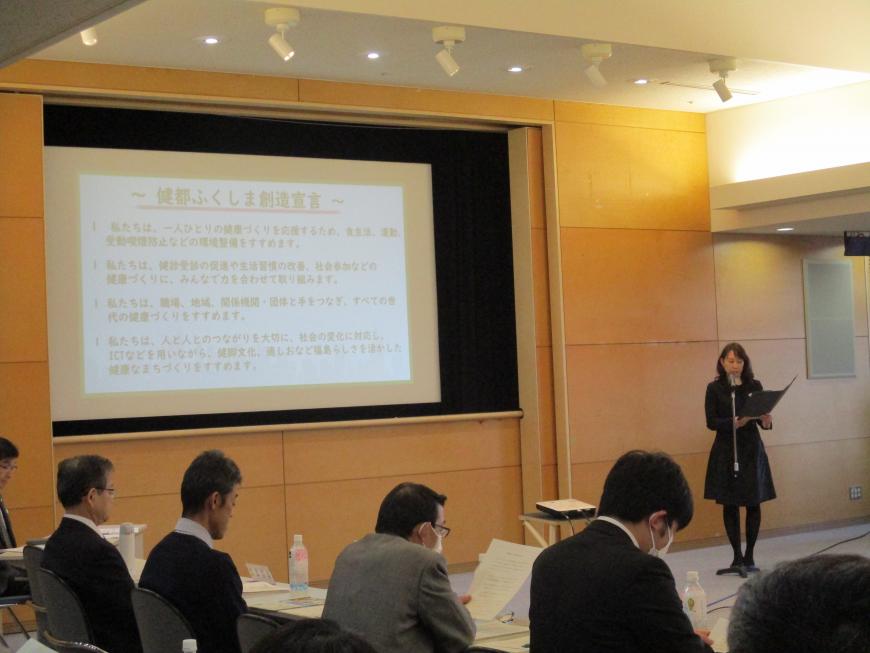 会場の大きなスクリーンに「健都ふくしま創造宣言」が映し出され、出席者が着席する中、スクリーンの横に立った女性が文書を読み上げている写真