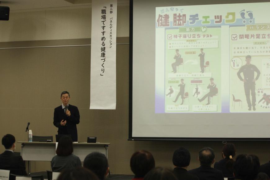 大きなスクリーンの左側に「第二部パネルディスカッション「職場ですすめる健康づくり」」と書かれた垂れ幕が下がっており、その手前に立ってマイクを持った男性が参加者席に向かって話をしている写真