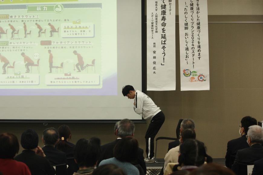 資料が映し出された大きなスクリーンの手前で、スクリーンの資料と同じ、椅子から立って膝と腰を曲げて前傾姿勢になるポーズをとっている男性の写真
