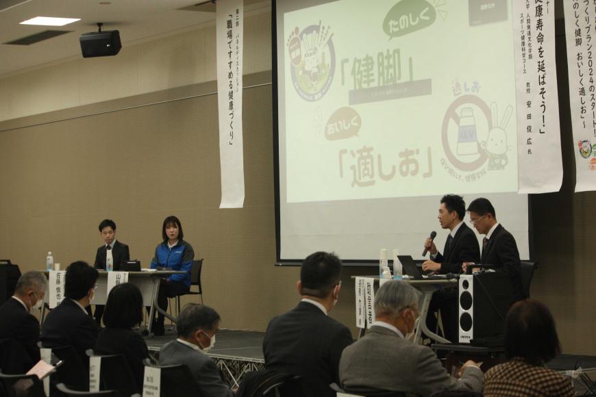 両側に複数の垂れ幕が下がった大きなスクリーンの手前に2人並んで座った男性のうち1人がマイクを持って話をしており、スクリーンを挟んで反対側の席に男女が座り、客席には参加者が座っている写真