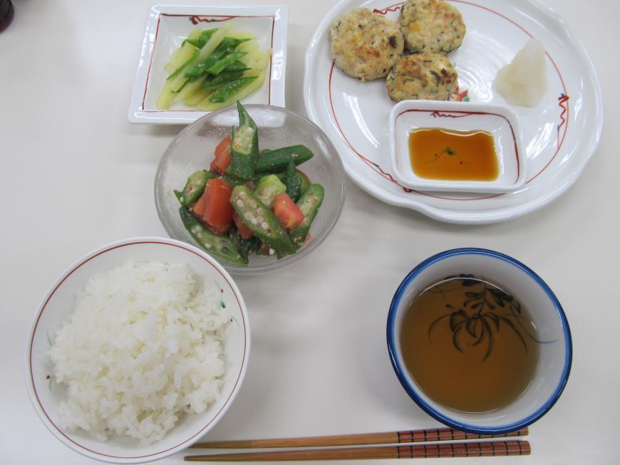 手前に箸とお茶、ごはん、中央にオクラとトマトの和え物、奥の左にじゃがいもと緑の野菜の小鉢、右にタレと大根おろしが添えられたつくね焼きが並んでいる写真