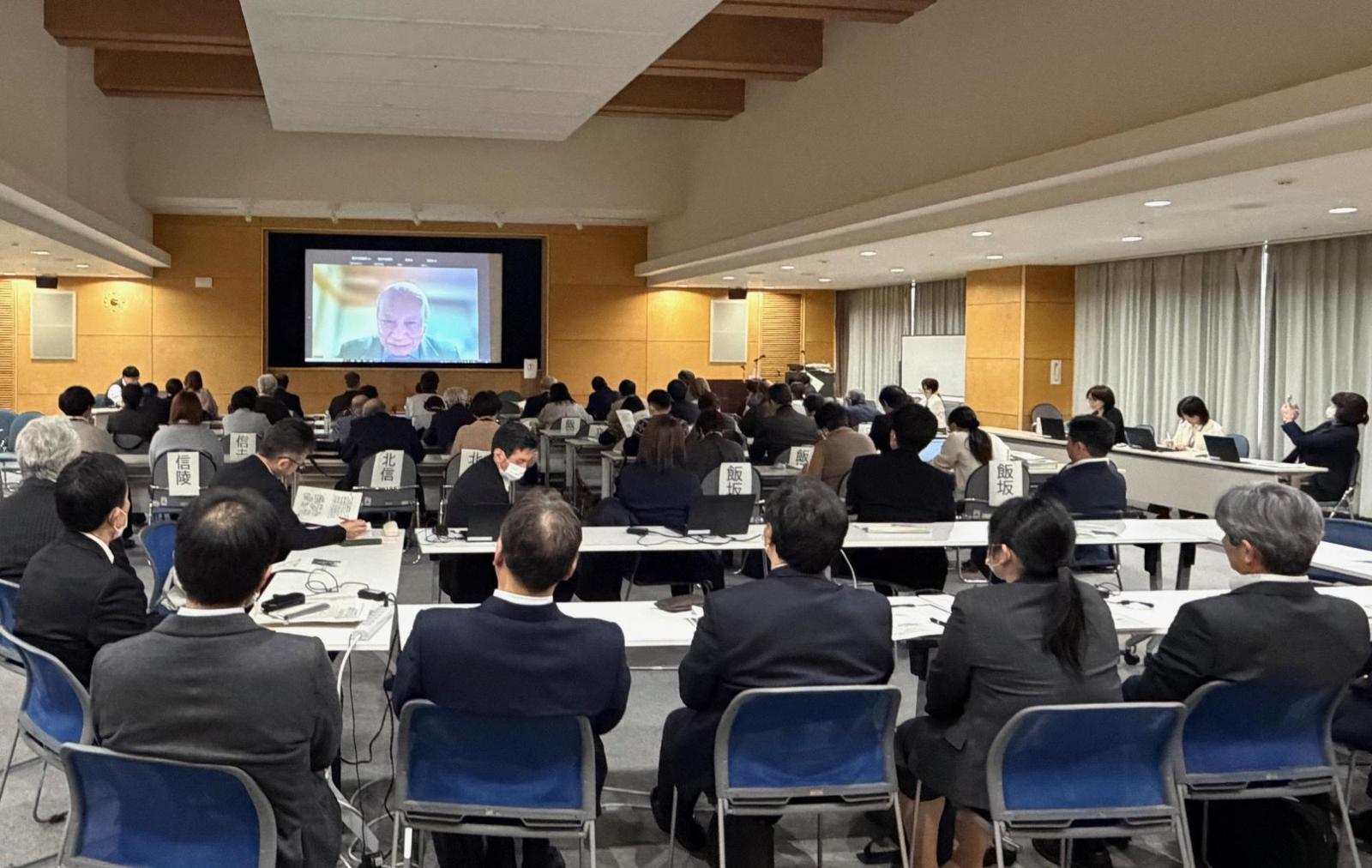 奥の壁に大きなスクリーンがあり、大勢の参加者がテーブルの席に座っている会場全体の写真