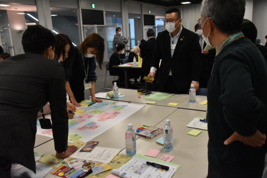 グループでの話し合いで、出てきた意見を付箋に書き出し、テーブルの周りに集まり話合っている参加者達の様子の写真