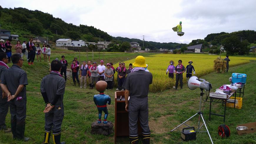 田んぼの前にレンガ色のタオルを首にかけた参加者達が集まり、前で関係者が挨拶をしている写真