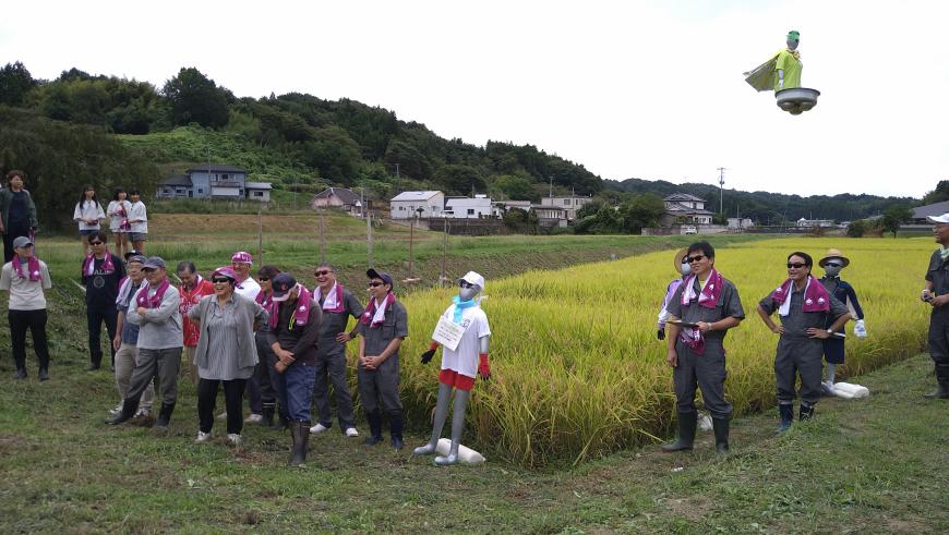 田んぼの前にレンガ色のタオルを首にかけた参加者達が集まっている写真
