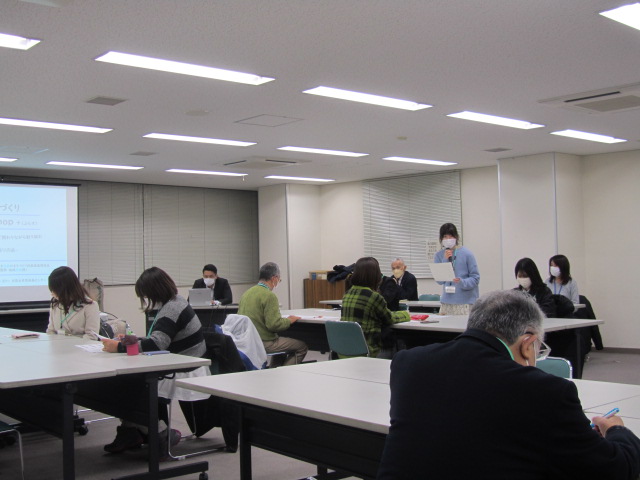 前方で女性が立って手元に資料を持って説明をするのを参加者達が聞いている写真
