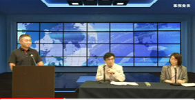 左側の演壇に男性が立っており、その横のマイクが設置されたテーブル席に男性と女性が座っているYouTube画面の写真