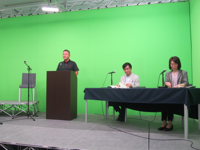 演壇に男性が立っており、横にマイクの設置されたテーブル席に男性と女性が座っている写真
