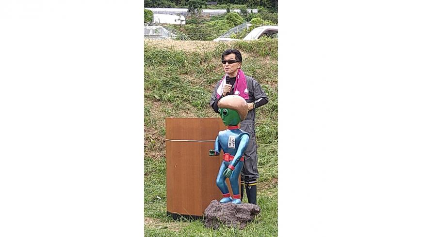 UFOの人形の後ろでマイクを持って話をする男性の写真