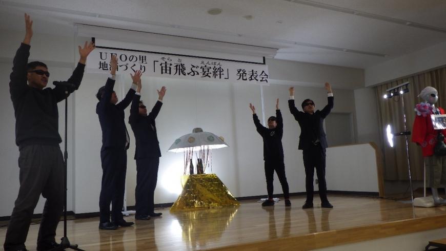 後ろに「UFOの里 新酒づくり「宙飛ぶ宴絆」発表会」と書かれスーツ姿にサングラスをした男性達が万歳をしている写真