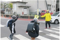ランドセルを背負った小学生が横断歩道を渡る様子を黄色の旗を持った女性が見守り活動をする様子の写真