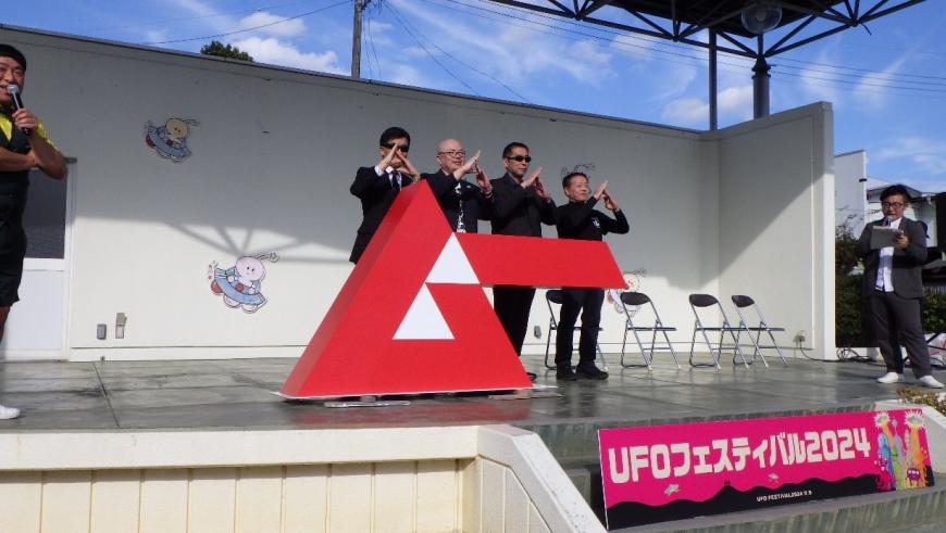 ステージ下に「「UFOフェスティバル2024」」と書かれ、ステージで黒いスーツを着た男性4人が同じポーズで立っている写真