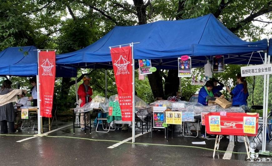 青いテントの出店に赤い旗が設置され、商品が並べられ青いTシャツを着た方が準備をする様子の写真