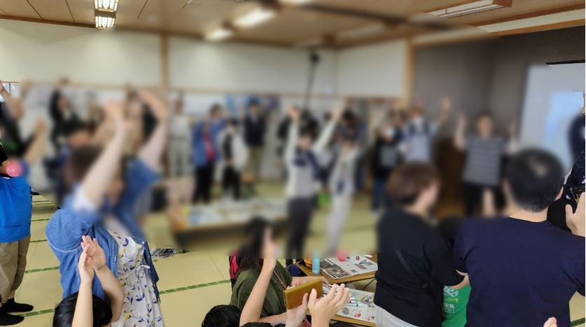 畳の敷かれた和室で参加者が両手を広げイベントに参加しているモザイクのかかった写真
