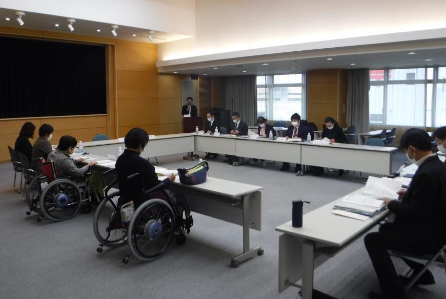 ロの字型に設置された長机の席に座った参加者たちが、前方右側に立つ男性の話を聞いている写真