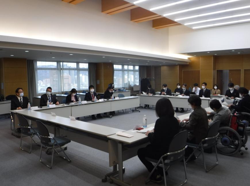 ロの字型に設置された長机の席に着席した参加者たちが資料に目を通している会議の様子の写真