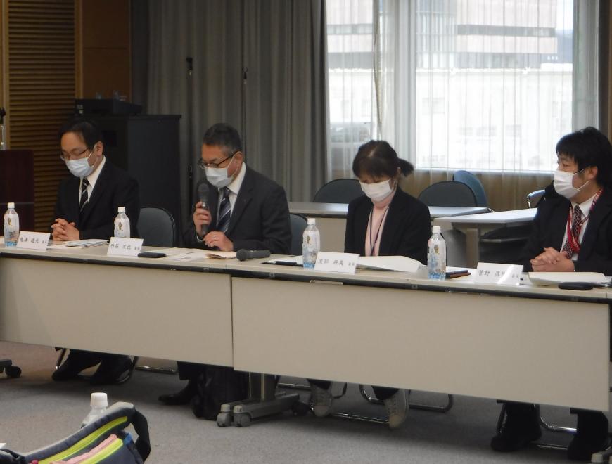 席札が置かれた席に参加者が座り、左から2番目に座った男性が右手にマイクを持ち話をしている写真