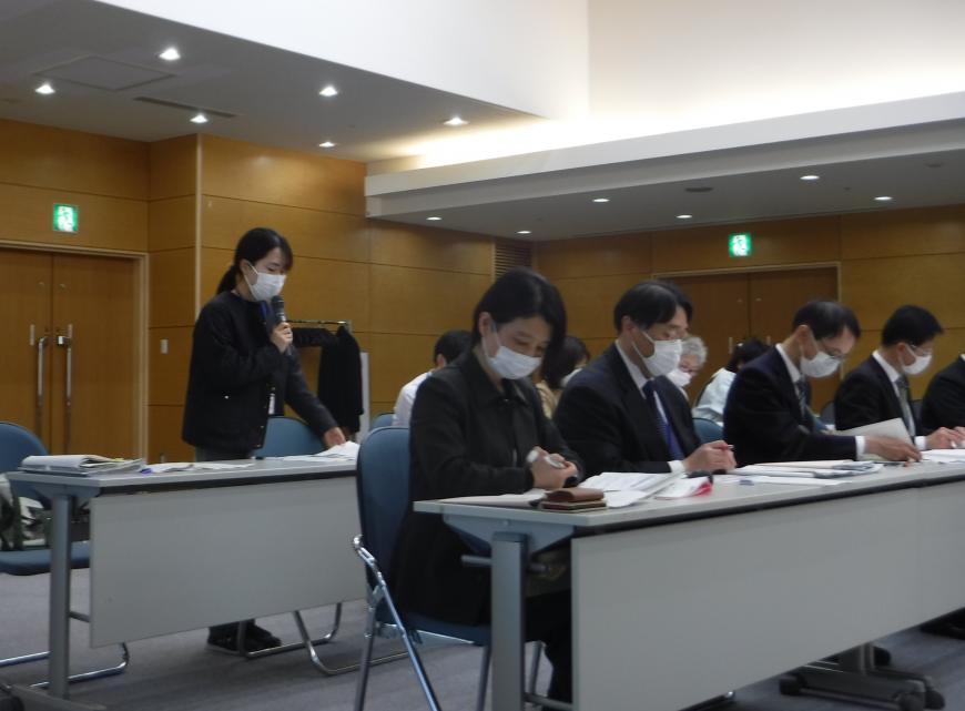 2列に設置された長机の席から立ち上がった女性が、右手でマイクを持ち発言している写真