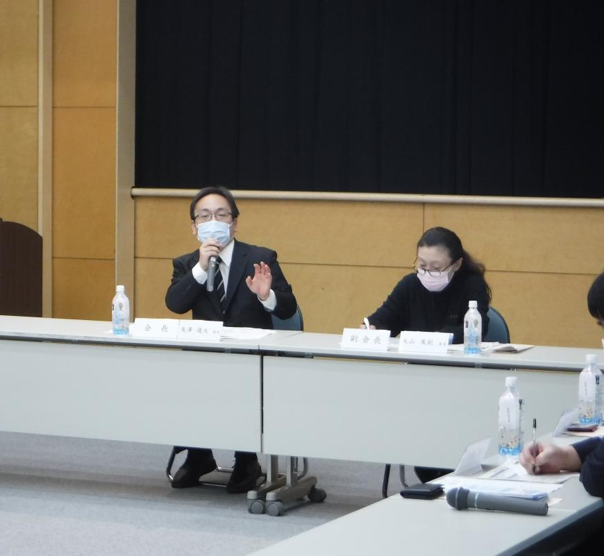 会長と副会長と書かれた席札が置かれた席に男性と女性が座り、鬼澤会長が右手にマイクを持ち話をしている写真