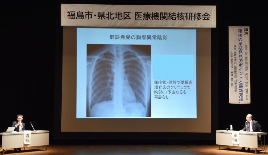 胸部レントゲン写真がステージ上の大きなスクリーンに映し出され、右側に「座長」と書かれた席に男性が座り、左側の「講師」と書かれた席の男性が説明を行っている情報交換会の様子の写真