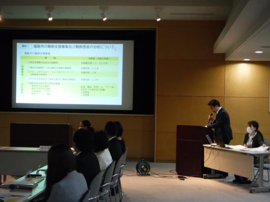 前方に資料が映し出されたスクリーンが設置され、右側に立つ男性がマイクを持ち説明を行っている写真