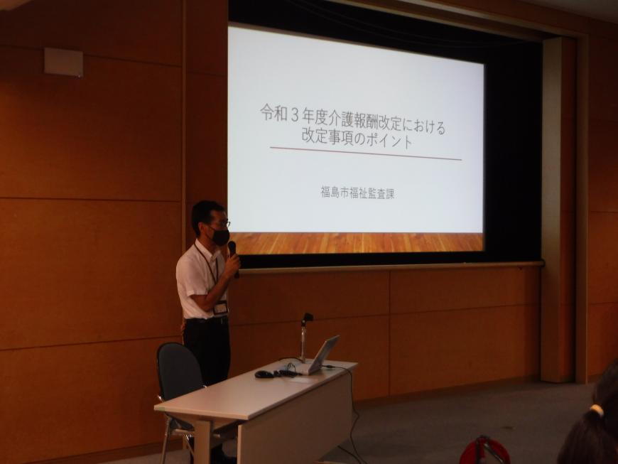 「令和3年度介護報酬改定における改定事項のポイント」と書かれた資料がスクリーンに映し出され、左側の席から立ち上がった男性がマイクを持ち話をしている写真