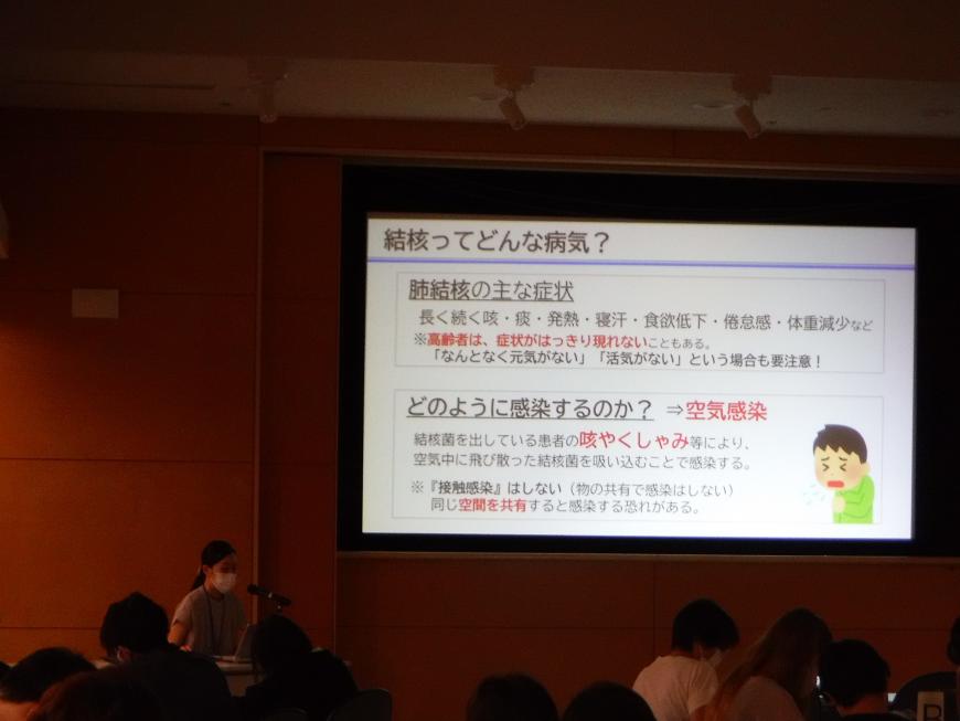 「結核ってどんな病気?」と書かれた資料がスクリーンに映し出され、左側の席に座った女性が話をしている写真