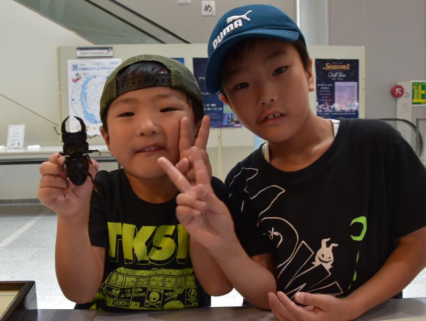 1人の男の子が片手でカブトムシを持ちもう一人の男の子とカメラに向かってピースサインをしている写真
