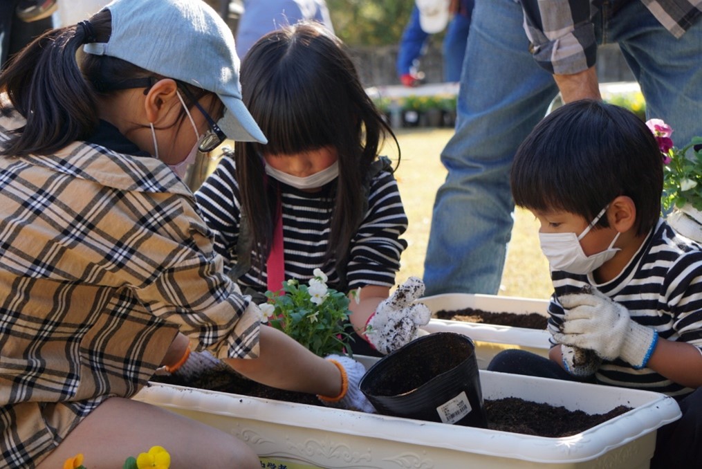 男の子と女の子の3名がプランターを囲み、1人の女の子がそっと花を植えようとしている写真