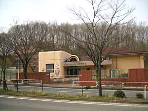 茶色の屋根とベージュの壁の平屋の建物が、茶色の壁の後ろにたっており、背後に林が広がる蓬莱第二保育所の外観写真