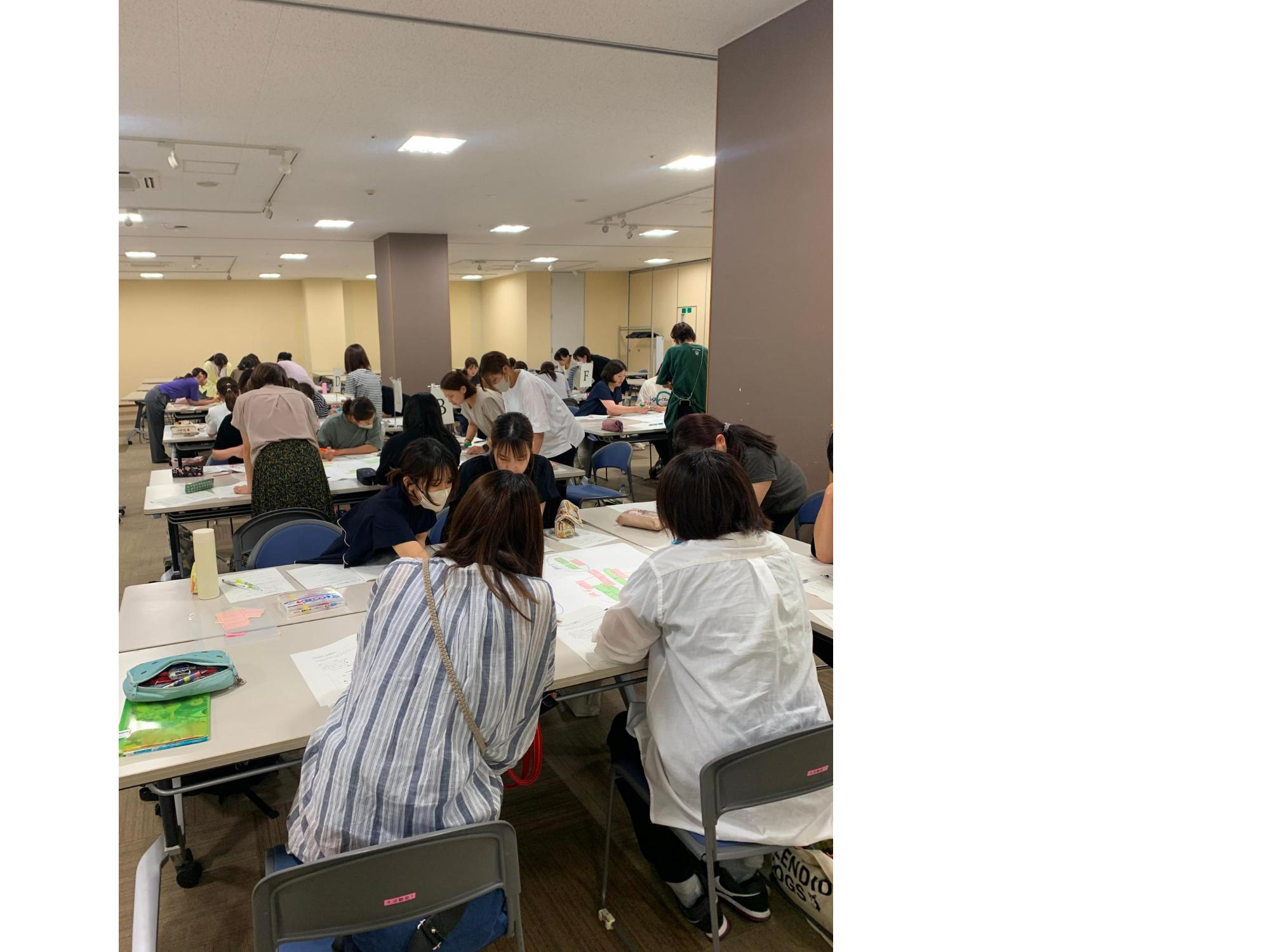 資料が映し出されたスクリーンやホワイトボードが設置された部屋で、グループに分かれてテーブルの席に座った参加者たちが話し合いをしている様子の写真