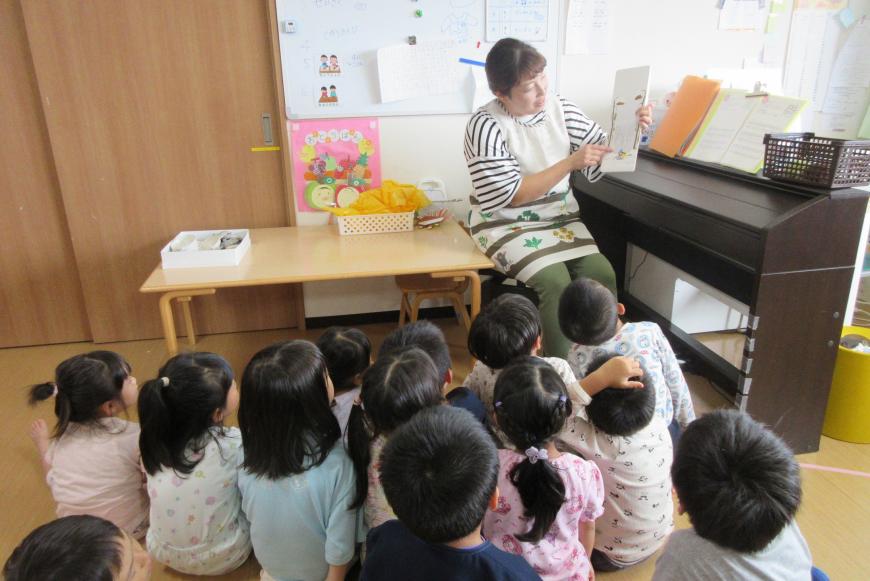 教室のピアノとテーブルの間に座った保育士の女性が、前に集まって床に座っている子どもたちに絵本の読み聞かせをしている写真