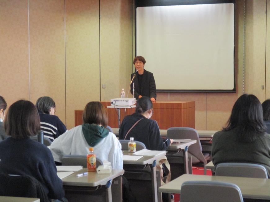 大きなスクリーンの前に立った女性が、演台に置かれたマイクで、席に着いた出席者たちに向かって話をしている写真