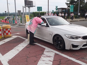 白い車の運転手にピンク色の法被を着た男性が記念品を配布している様子の写真