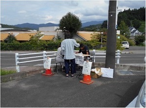 歩道の隅に設置された簡易テーブルを挟んだ椅子に2人座りアンケートを記入している様子の写真