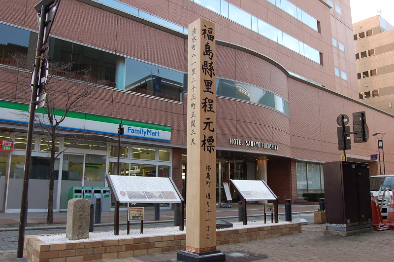 町中に設置された2つの説明看板の左側に福島市道路元標、前に福島縣里程元標が設置された写真