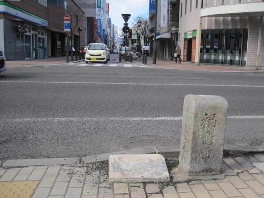 大きな道路を背に歩道に立つ福島市道路元標を写した写真