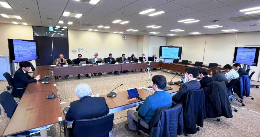 室内の四隅に大型モニターが設置され、ロの字型に設置した長机の席に関係者が座っている会議の様子の写真