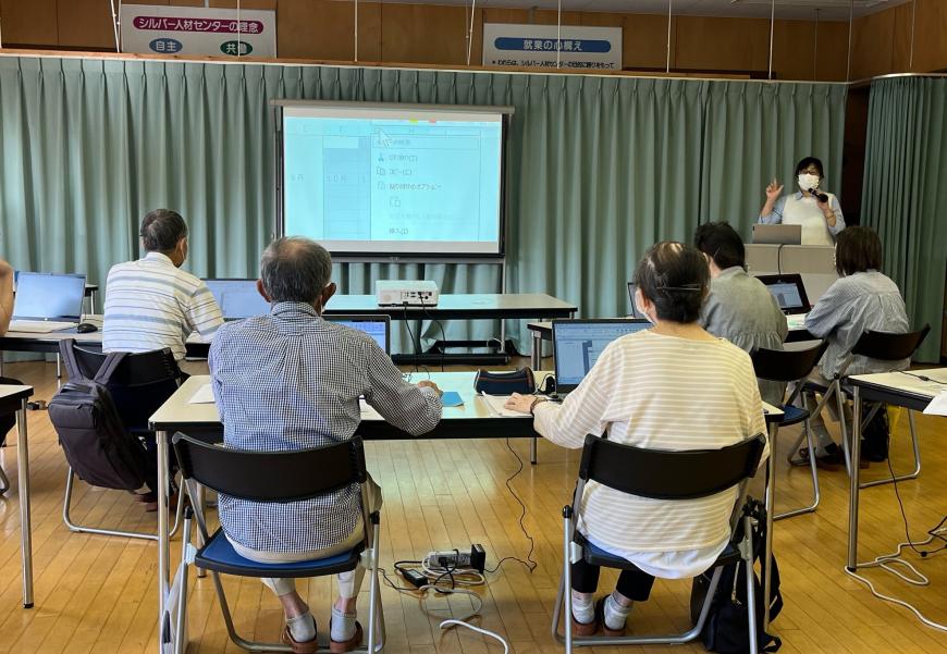長机の席に座った参加者たちが、前方に設置されたスクリーンを見ながら、右側に立っている女性の説明を聞いている写真