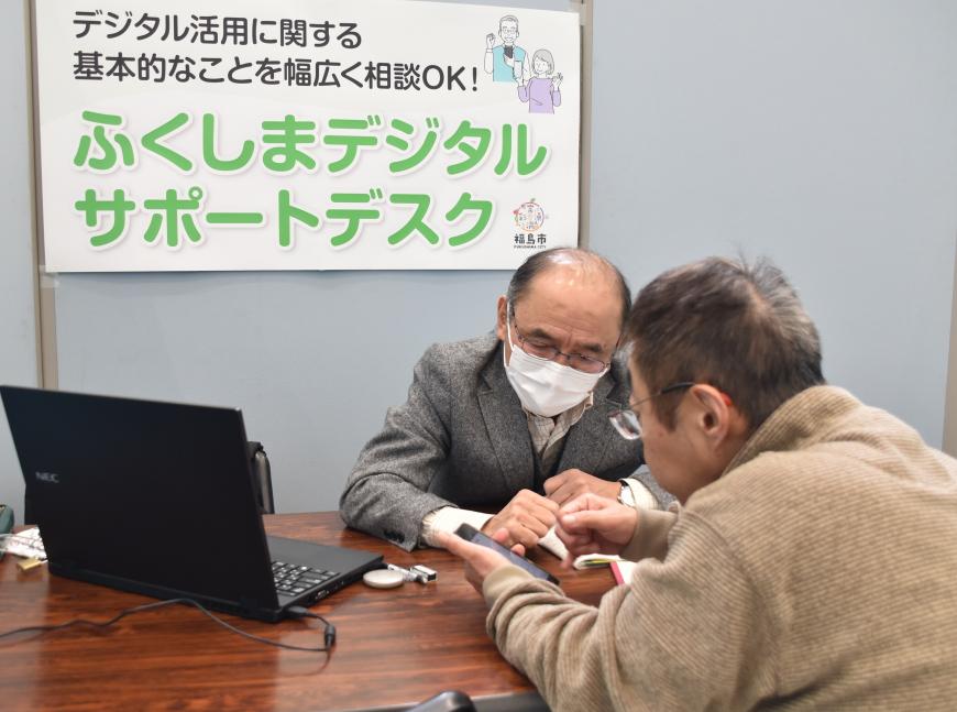 ふくしまデジタルサポートデスクと書かれた看板の前で男性担当者にスマートフォンの画面を見せて相談している男性の写真