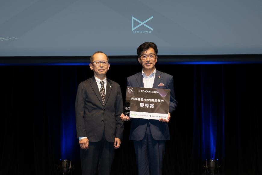 行政機関・公的機関部門優秀賞と書かれた盾を両手で持つ市長と関係者の男性が並んでいる表彰式の様子の写真