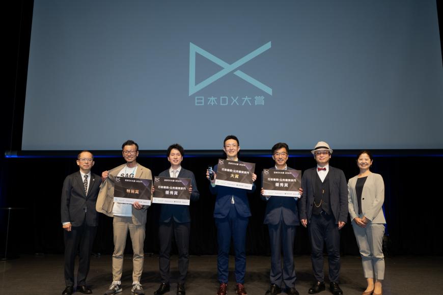 日本DX大賞のロゴマークが表示された背景をバックに、盾を持った市長を含め4名と3名の関係者が横一列に並んでいる写真