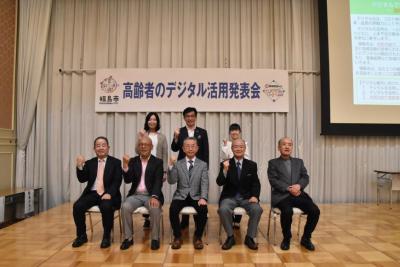 「高齢者のデジタル活用発表会」と掲げられたステージ上の前列の椅子に5名の男性、後列に市長と2名の女性がガッツポーズをしている集合写真