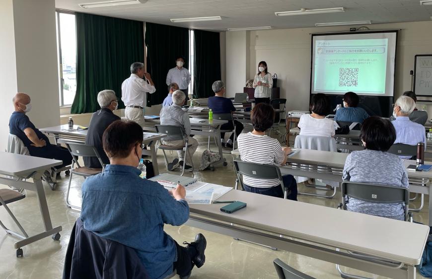 室内前方に設置されたスクリーンに資料が映し出され、長机の席に2人ずつ座った参加者が聞いているなか、左側の前から2番目の席の男性が立ち上がり話をしている研修の様子の写真