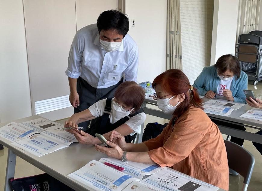 資料が置かれた長机の席に隣り合わせに座った2名の女性が、お互いスマートフォンを持ち教え合っている様子を後ろに立つ男性講師が見ている写真