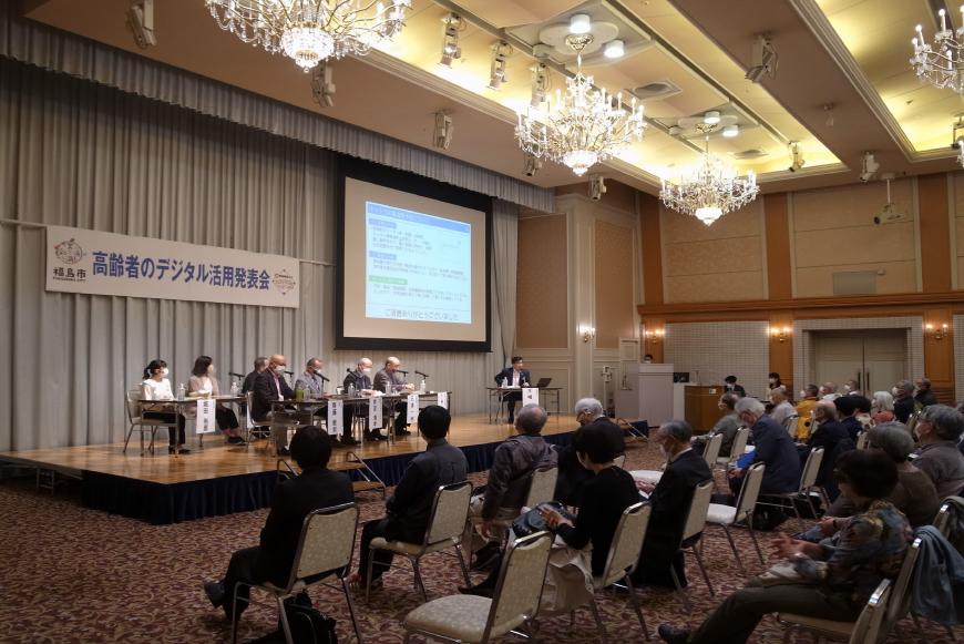 天井にシャンデリアが設置された会場のステージ上の席に関係者が座り、会場に設置された椅子に参加者が座って話を聞いている写真