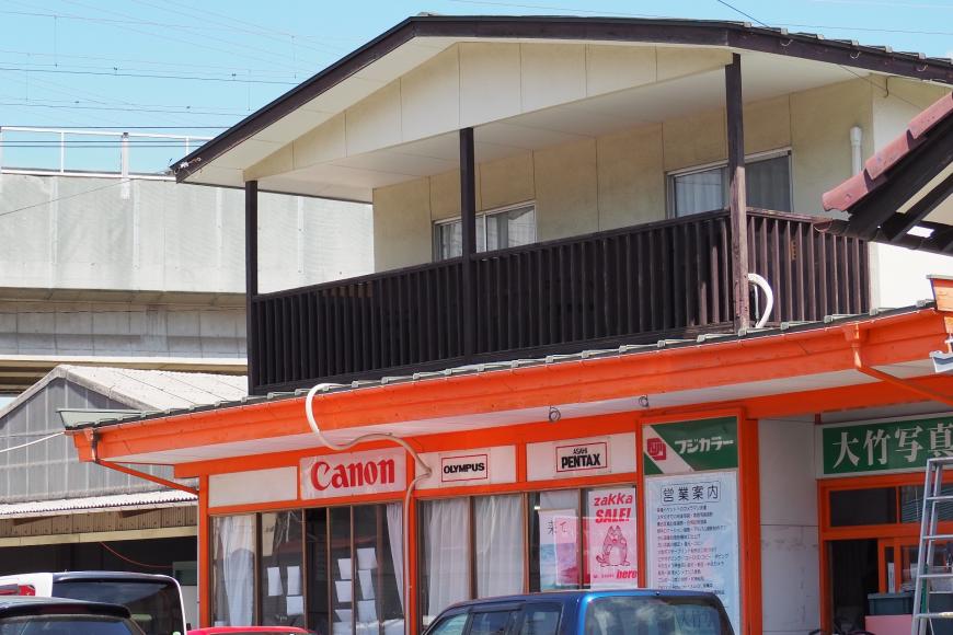 1階はオレンジ色の枠にガラス張りの店舗、2階は白壁のこげ茶色の枠の建物外観の写真
